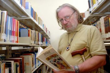 Homem de cabelos, barba e bigode brancos segurando um livro em maio a estantes da biblioteca